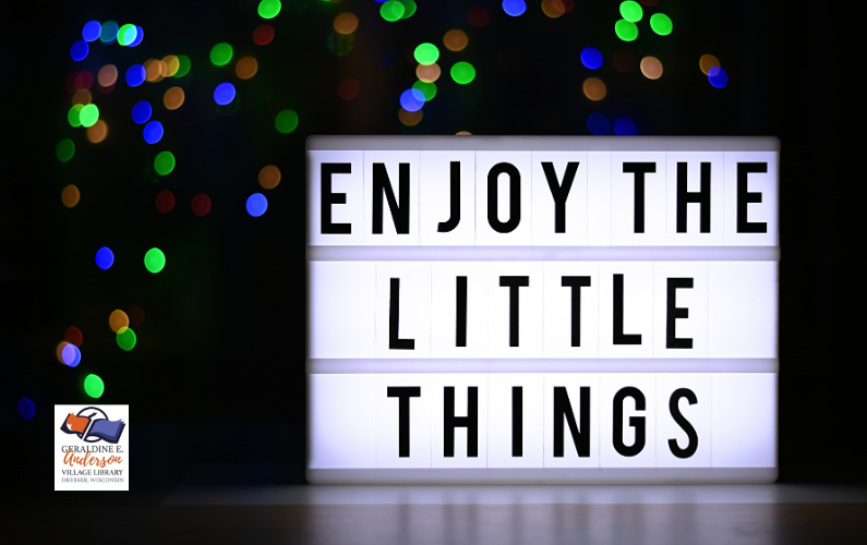 Sign with text saying "Enjoy the little things" under holiday lights