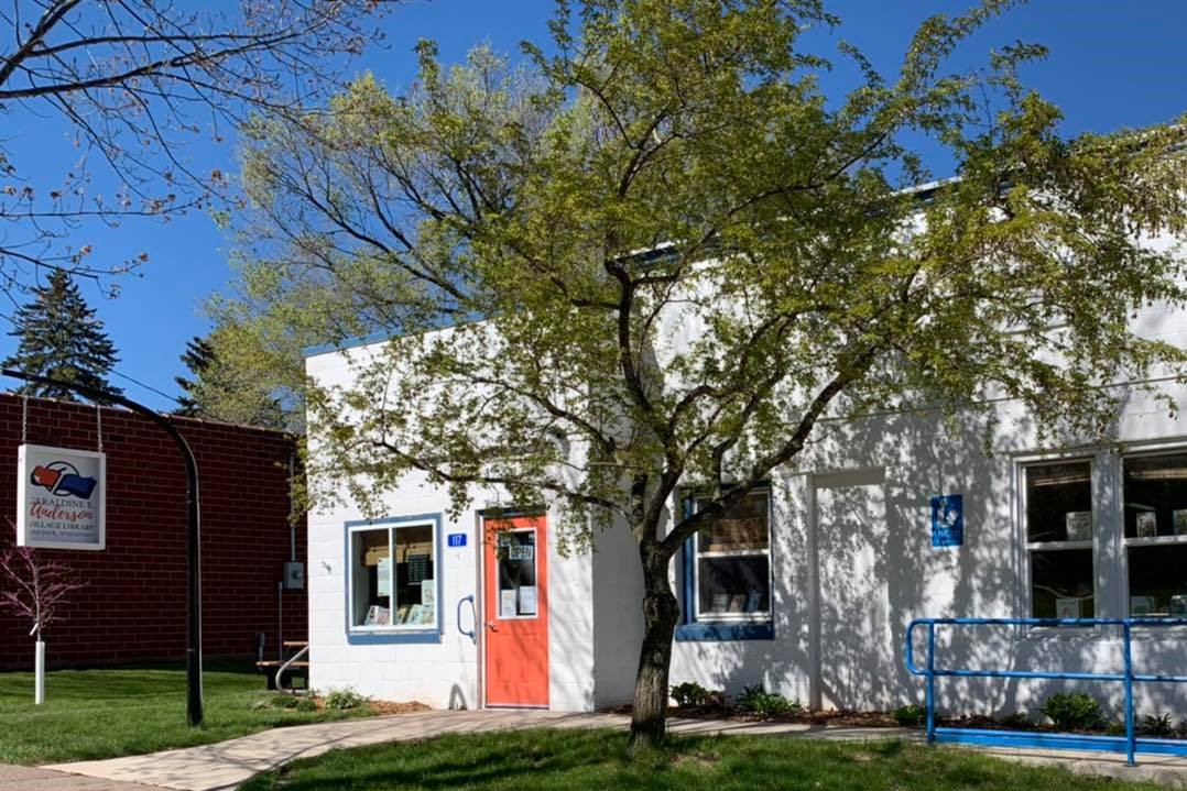 Small white library with an orange door, blue trim, and a sign out front under a leafy tree.