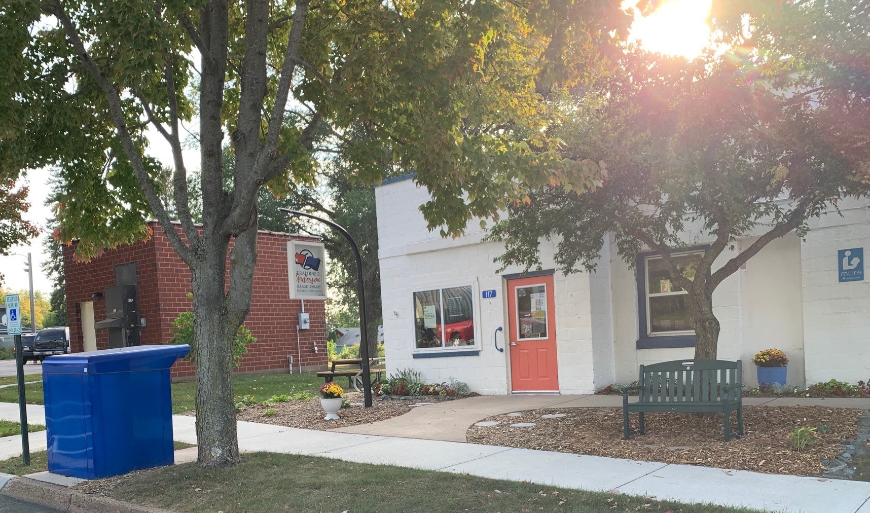 Dresser Library: White building with orange door with blue book return and two trees in the foreground with sunset shining through the leaves.