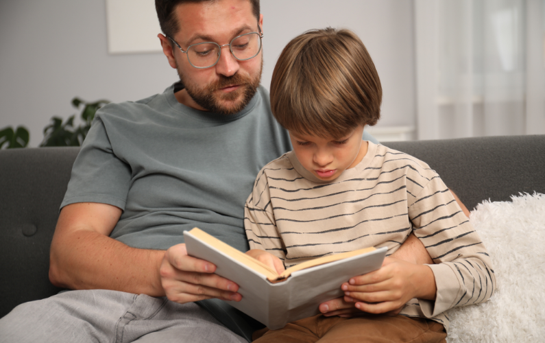 A man wearing glasses and a grey t-shirt sits on a sofa, holding a book open with a child sitting in front of him, also looking at the book.