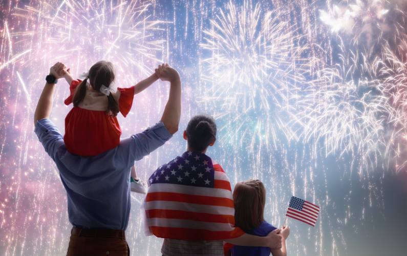 A family watches fireworks. One child is on someone's shoulders, another child holds an American flag, and another person is wrapped in an American flag.