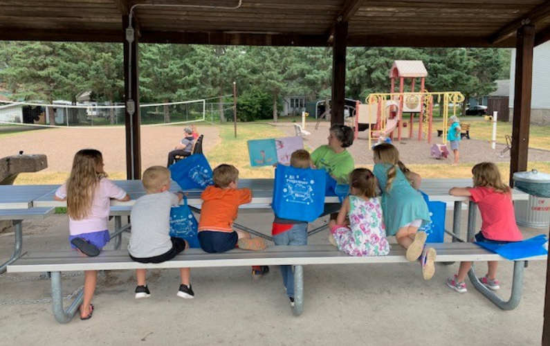 A group of children sit at a picnic table under a covered pavilion, listening to a story with a playground and volleyball court in the background. Several blue tote bags are on the table in front of the children.