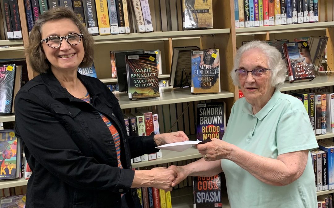 Two women standing in front of a library book shelf exchanging an envelope.