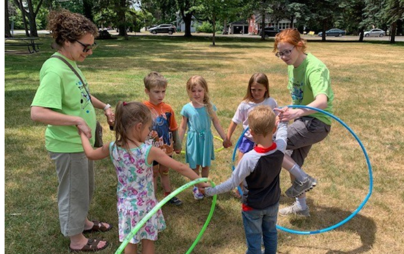 Two adults and several children hold hands in a circle in a grassy park, with some children holding hula hoops.